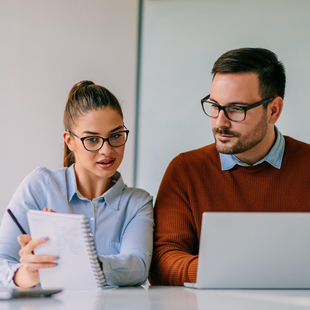 Zwei Menschen. Frau und Mann. Mann und Frau beraten sich. Frau hält Notizblock. Mann sitzt am Laptop und schaut auf den Notizblock.
