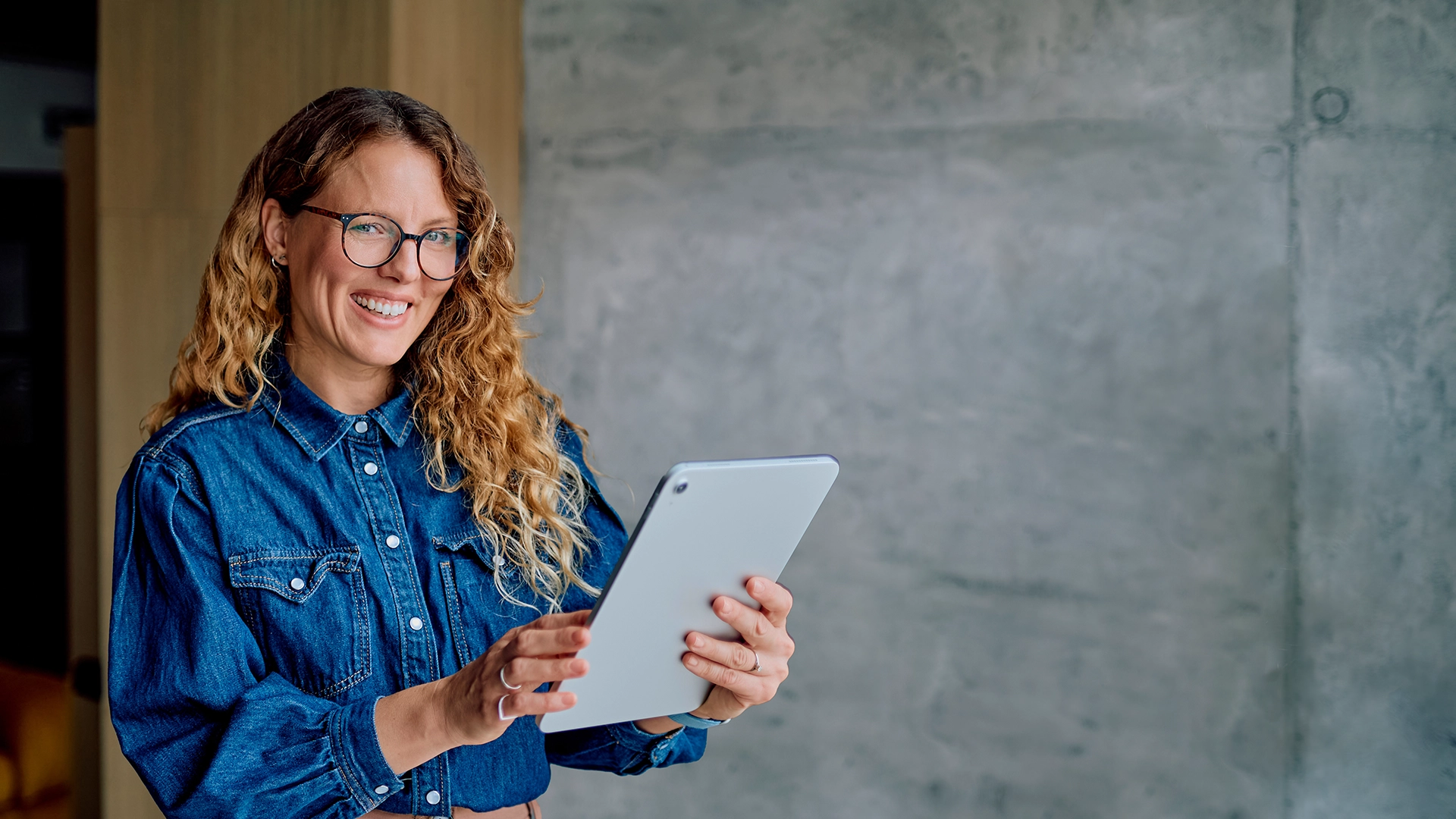 Eine Fachberaterin für Servicemanagement steht mit einem Tablet in der Hand vor einer Betonwand und schaut in die Kamera.